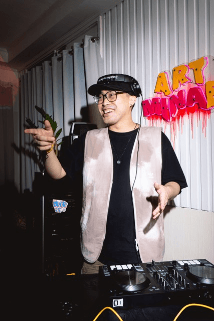 DJ pointing at the camera behind turntables, with graffiti-style "ART" lettering on the wall behind him.