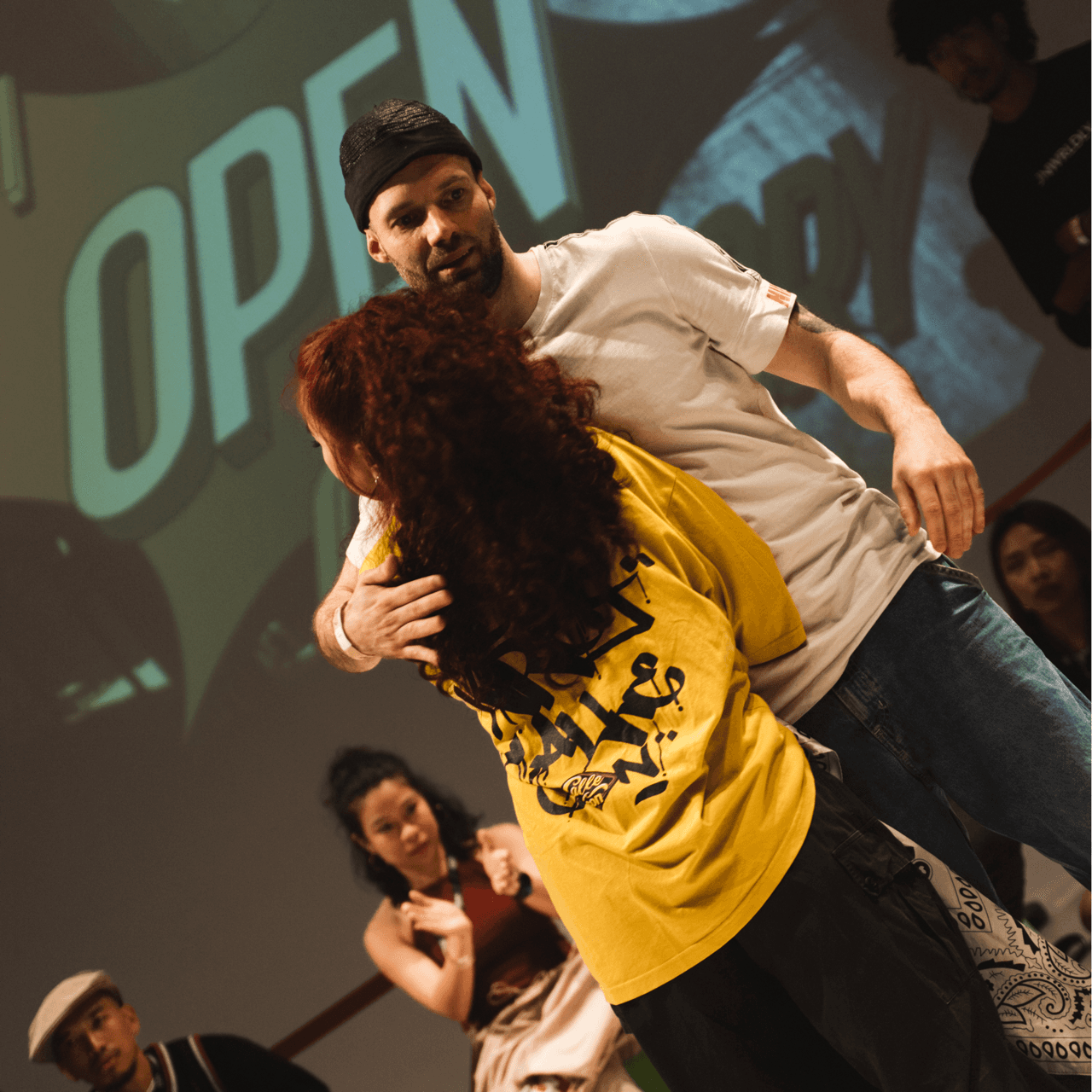 Man hugging a woman with curly red hair on stage in front of graffiti-style "OPEN" text.