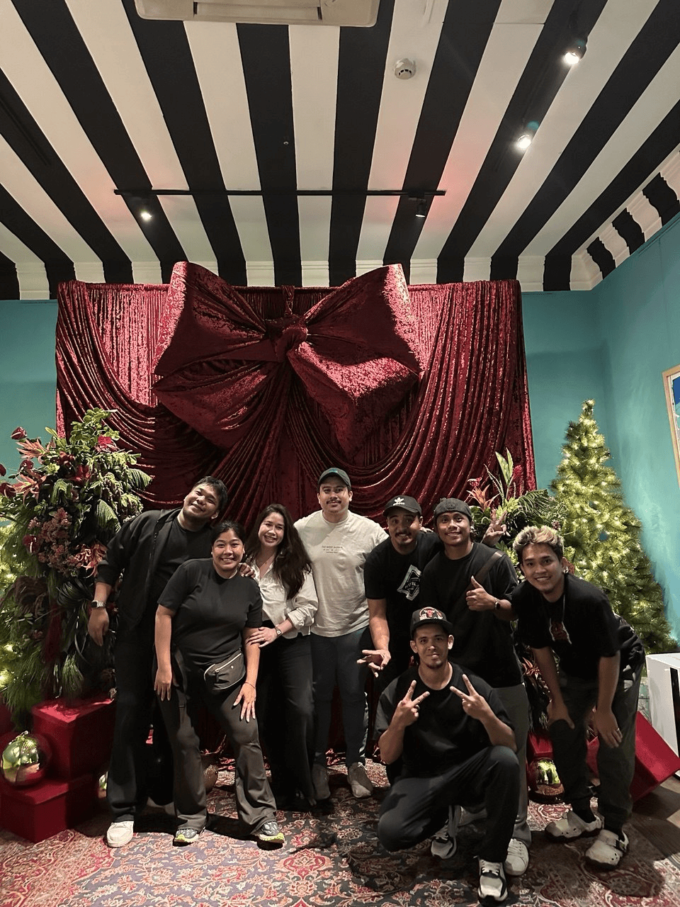 Group photo in front of a giant velvet gift-bow backdrop and Christmas tree, striped ceiling above.