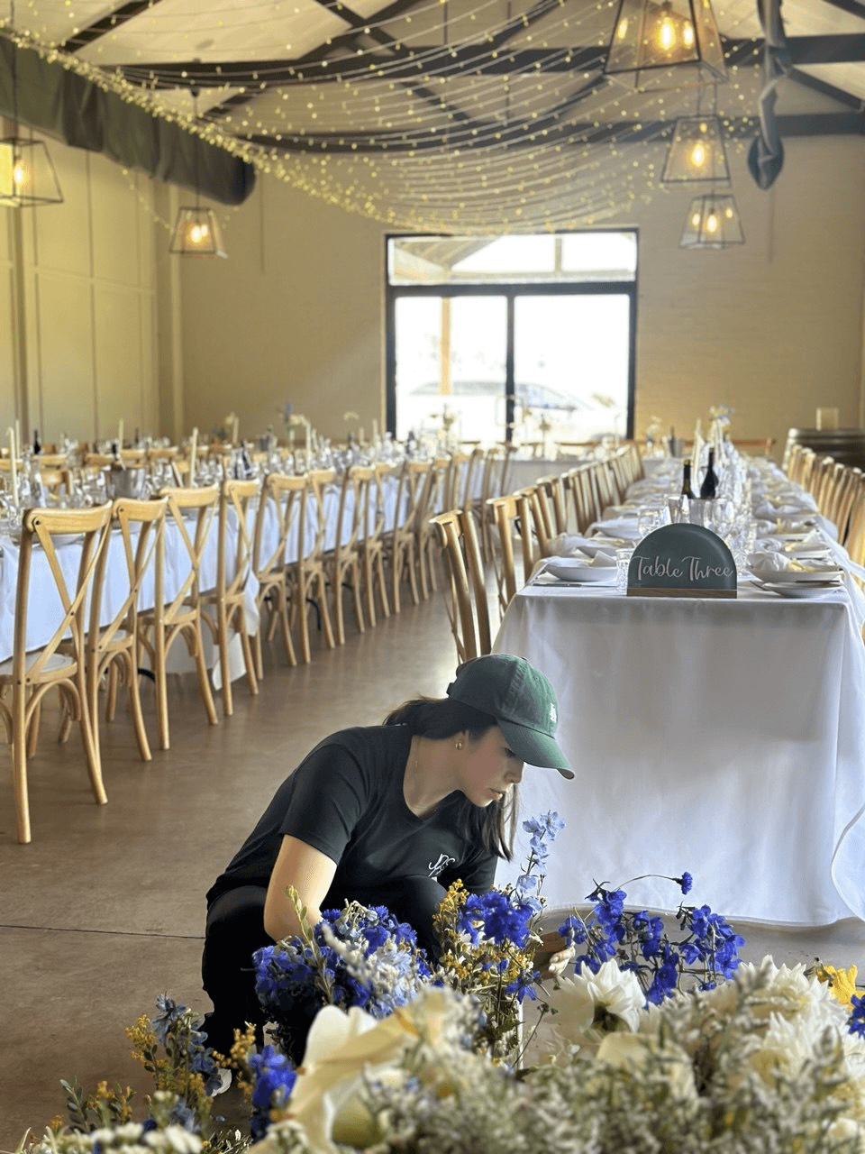 Woman arranging blue and white flowers beside long banquet tables set for a wedding reception, string lights overhead.