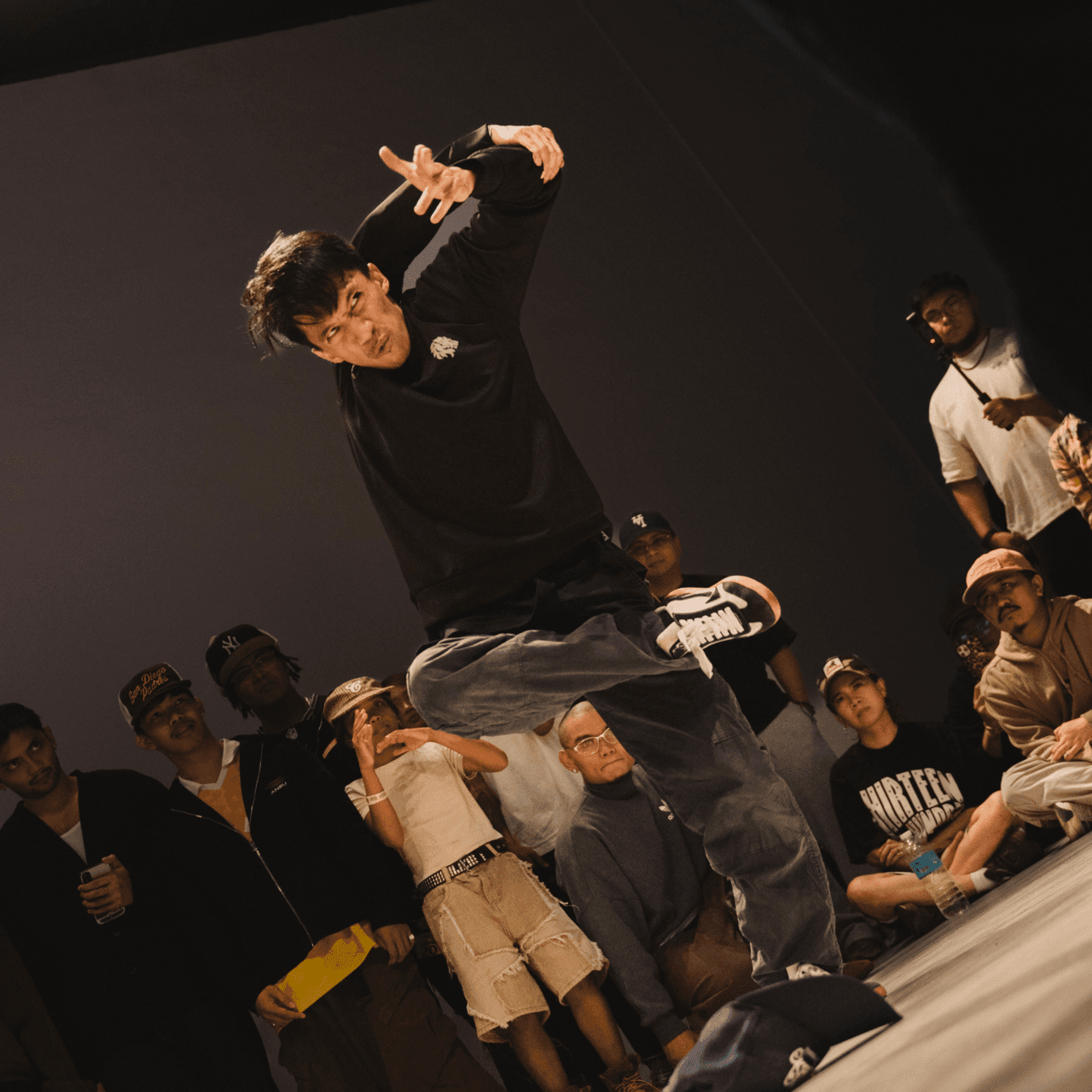 Breakdancer mid-freeze on one hand, legs extended, circle of onlookers watching from the floor.