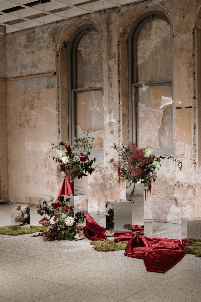 Floral arrangements on mirrored pedestals with red fabric and moss, set against a weathered, arched wall.