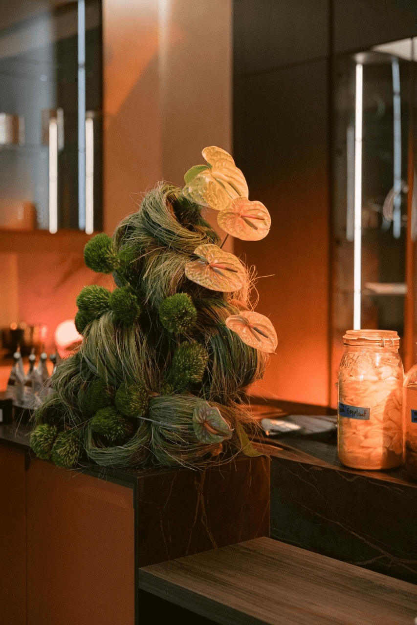 Cone-shaped floral arrangement of green pom-pom mums and pale anthuriums on a bar counter, lit in warm orange light.