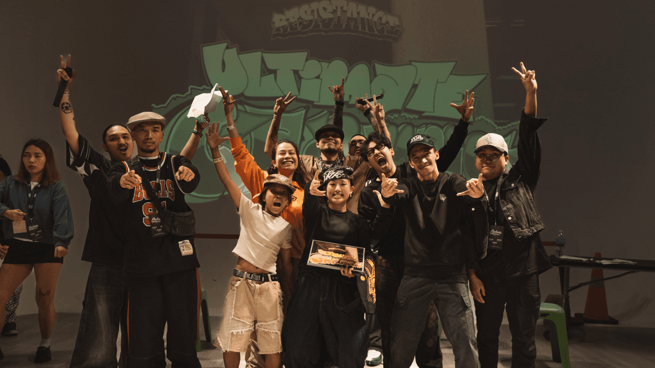 Group photo on stage, arms raised and throwing peace signs, in front of a graffiti-style "Ultimate" backdrop.