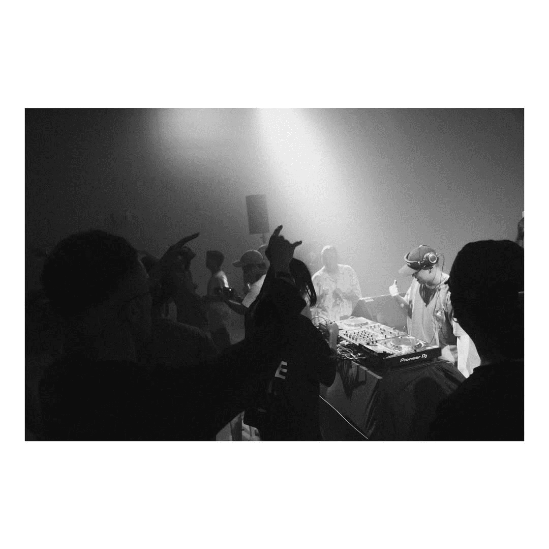 Black-and-white shot of a DJ at Pioneer turntables, crowd dancing in silhouette under a hazy spotlight.