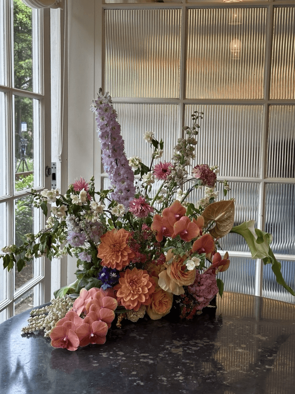 Large mixed floral arrangement with dahlias, delphinium, and orchids on a table by fluted glass windows.