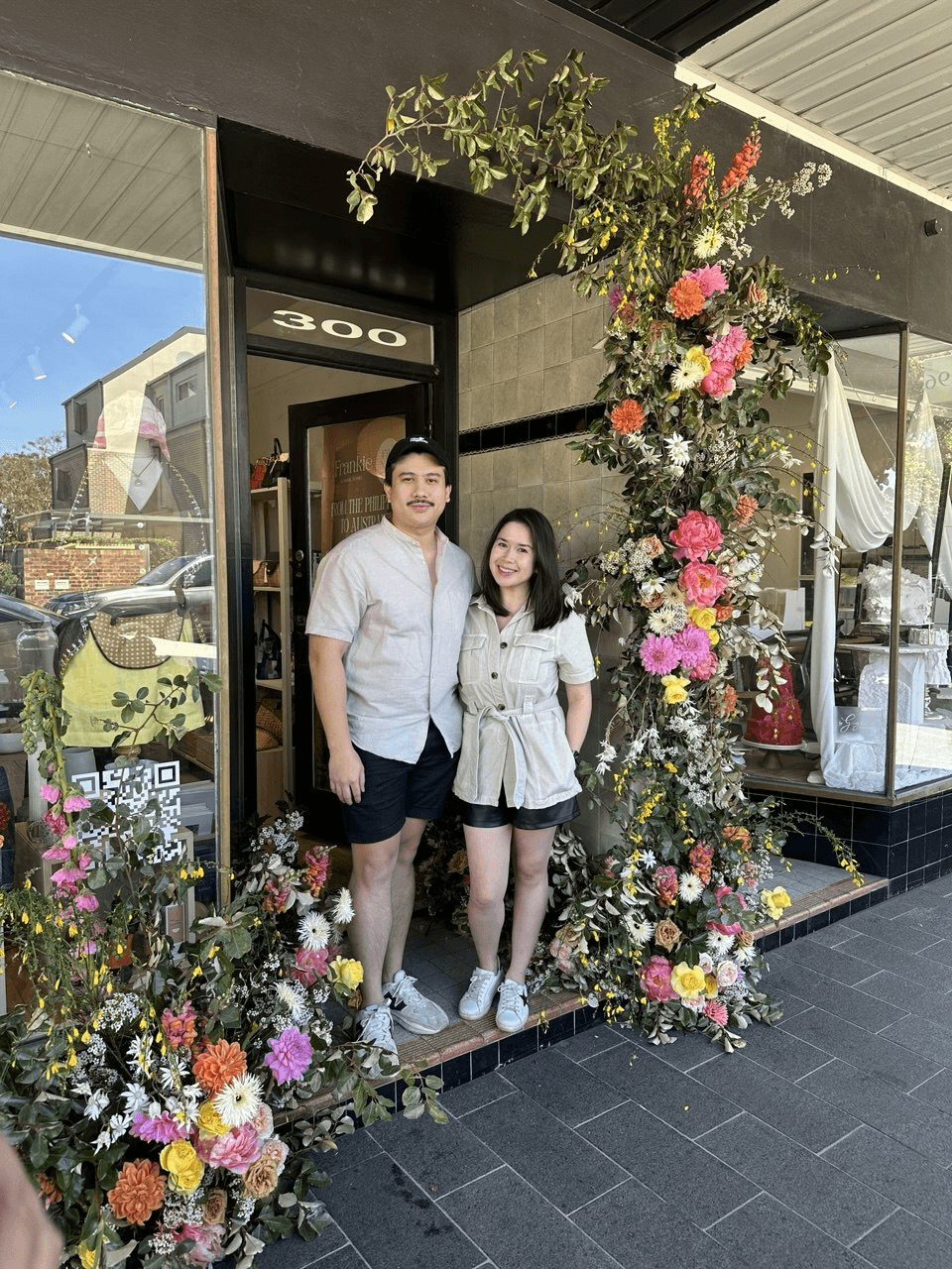 Couple standing in a shop doorway framed by a large floral display, storefront numbered 300.